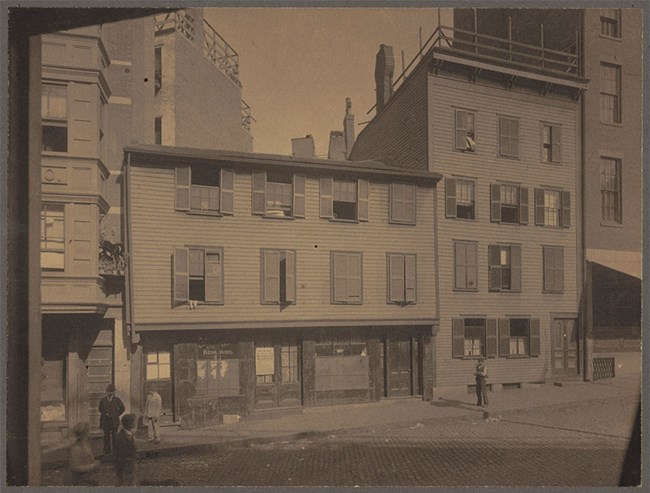 Black and white photograph of the Paul Revere House as a three story building serving as a tenement and storefront.