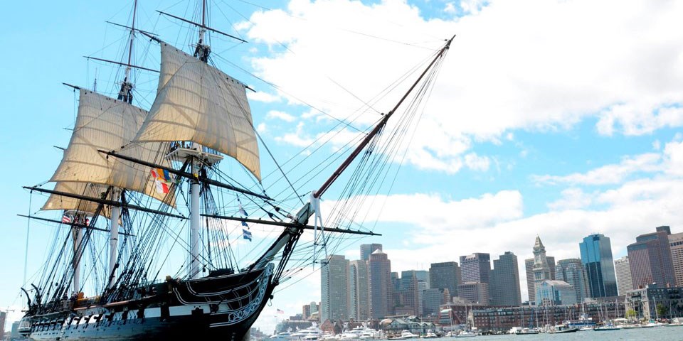 USS Constitution with sails rigged in Boston Harbor. The Boston skyline is in the background
