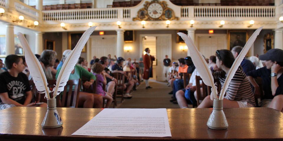 Quill pens in an inkwell flanking a document on a table at the head of the Great Hall. In the background the Great Hall of Faneuil Hall is full of seated participants while a man dressed in colonial garb stands up to address the meeting.