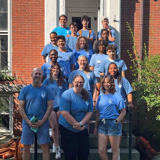 Multiple rows of youth stand in uniform blue tshirts in front of a brick building's entrance.