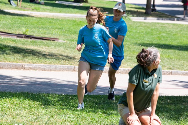 Two kids in blue shirts and shorts run, one chasing the other in the grass as someone in a green shirt kneels on the ground.
