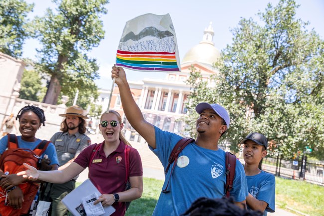 Youth in a blue tshirt holds up a flag with silhouettes standing above a rainbow path. Others, including a park ranger, stand around.