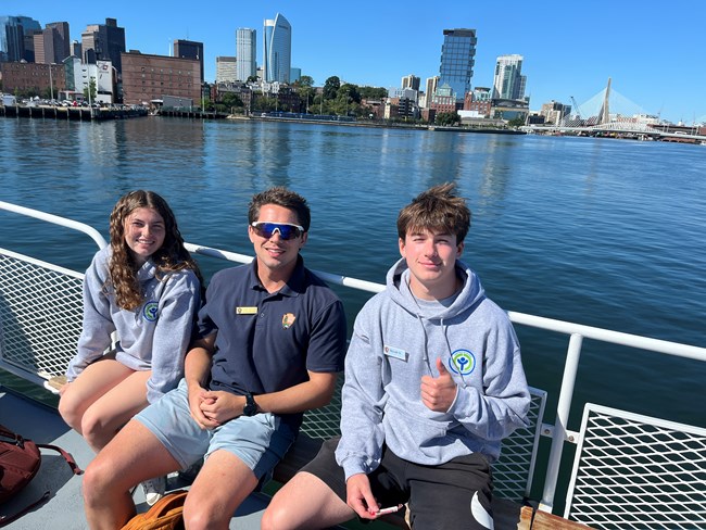 A group of three youth sit on a boat in the harbor with a city skyline behind them
