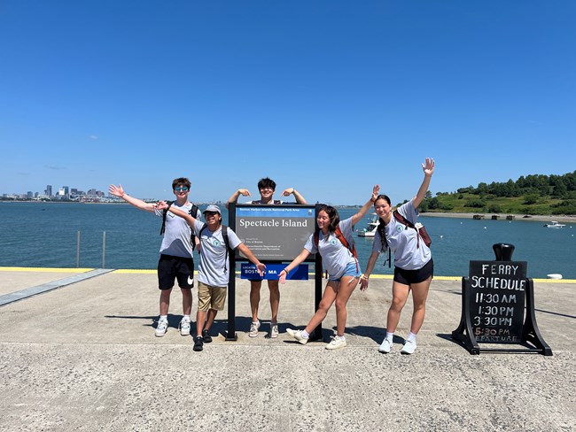 A group of youth pose in front of a sign in front of Boston harbor