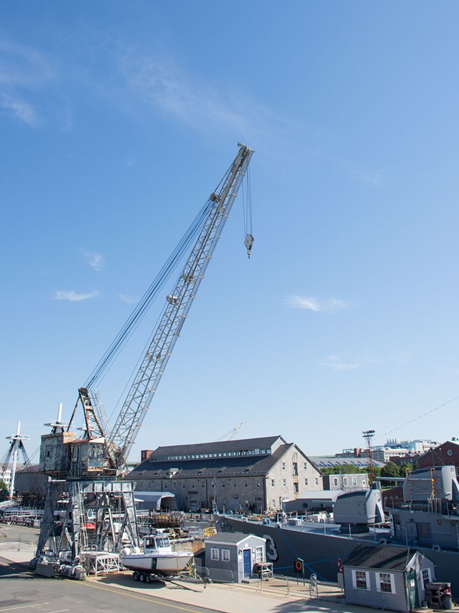 A steel portal crane towers over the bow of a docked warship. Equipment and tool sheds are nearby on the pier. A granite warehouse stands in the background.