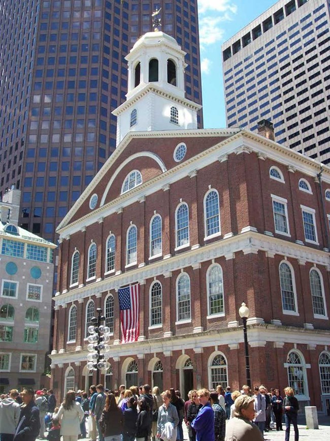Faneuil Hall is a four-story brick building with arched windows. The top of the hall has a while cupola with a gold dome and a grasshopper weathervane. Crowds of people pass in front of the hall.