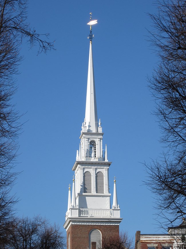 Belfry of Old North Church. The white belfry sits atop a brick base and an arched window is above. Atop the belfry is a tall spire with a golden weathervane.