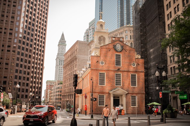 an older 3 story brick building with a white steeple amidst skyscrapers.