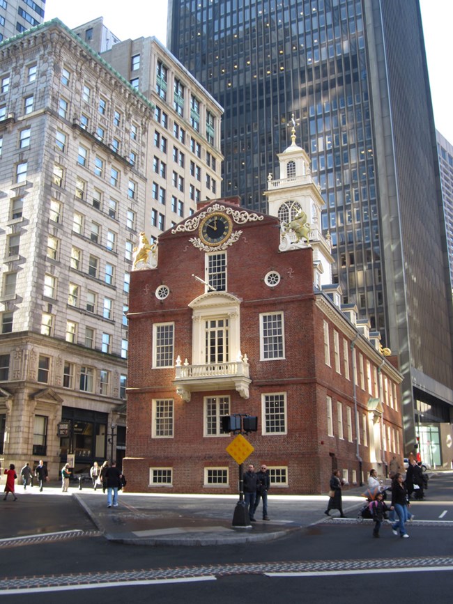 Old State House is a three story brick structure with dormer windows along the roof. A second floor balcony overlooks a main intersection of the city. A clock face is above the balcony and a cupola adorns the top of the building at the center.