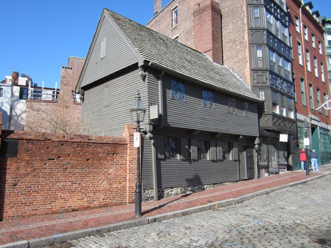 The Revere House is a two story wooden building painted gray. The high pitched roof is cedar shingle with a chimney on the right end of the house. The windows are panes of diamond-shaped glass. Shutters are only on the first floor windows.