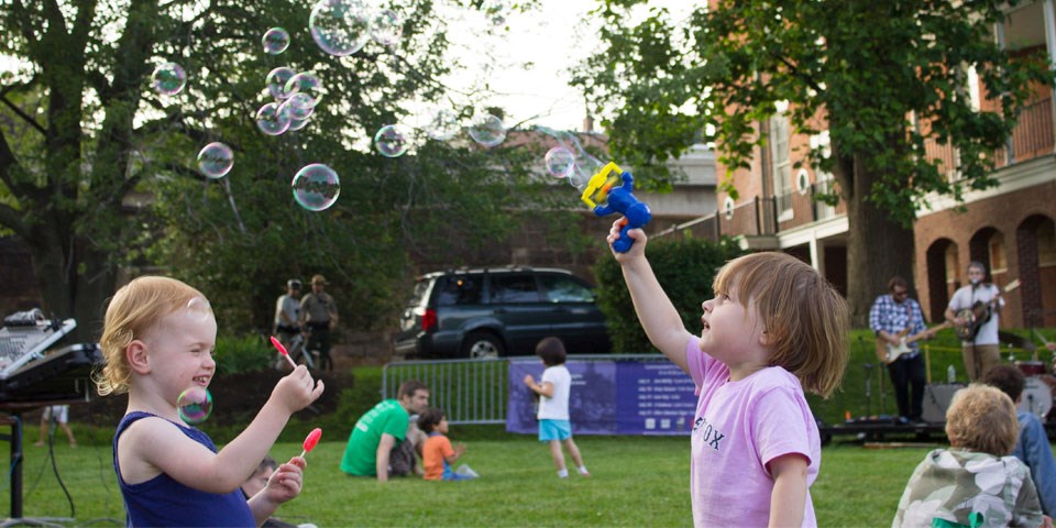 Two children play with bubbles on a lawn. Musicians play on a stage in background as crowds watch.