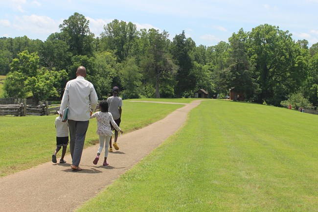A man and three children walk along a reddish earthen path with a small cluster of rustic buildings in the distance.