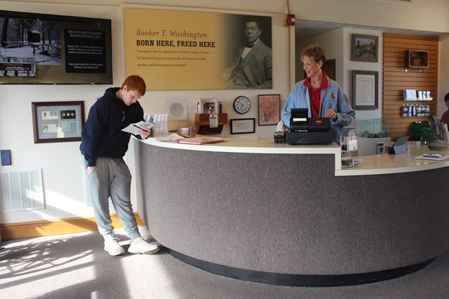A photograph of a visitor center desk with a woman behind cash register and a visitor reading some information.