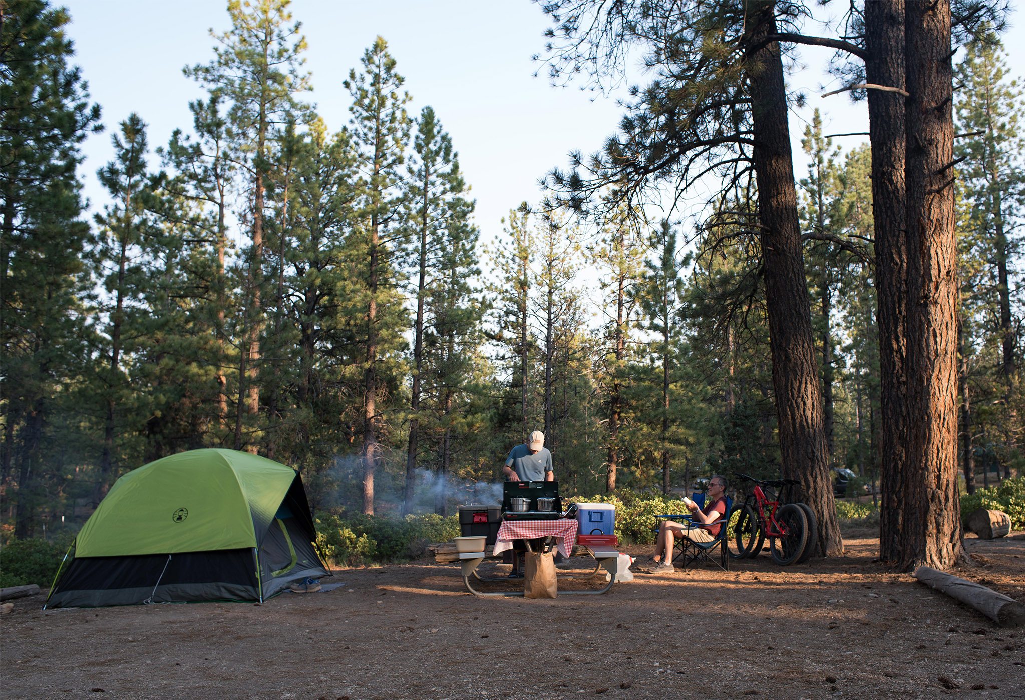 A man stands at a picnic table cooking at a stove near a tent while another sits in a camping chair at a forested campsite