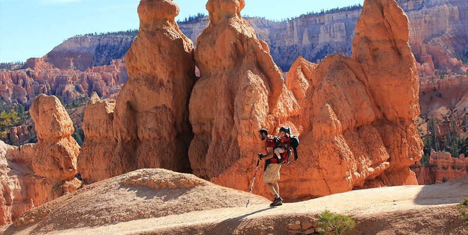 Man carries child on back along trail surrounded by red rock spires