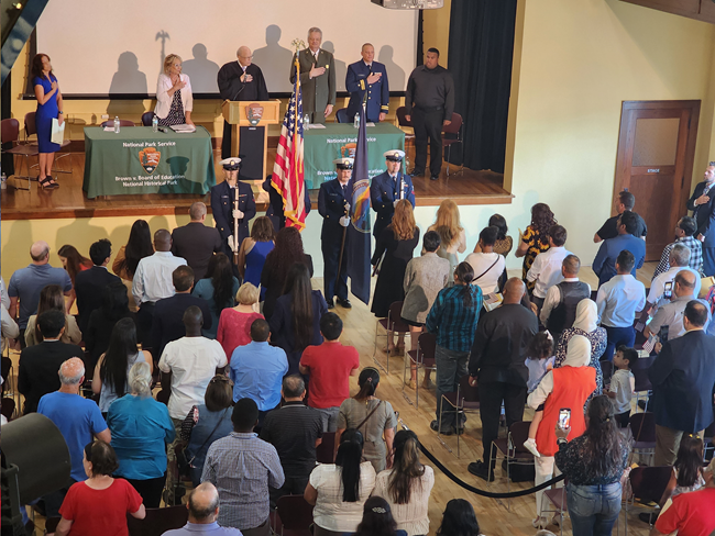 Attendees of a naturalization ceremony stand and place their hands over their hearts in an auditorium with wooden floor.