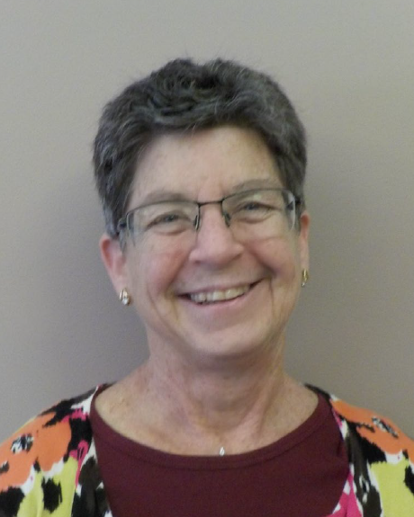 Head shot of a middle aged woman with short black and white hair, modern glasses gold earrings and colorful floral shirt.