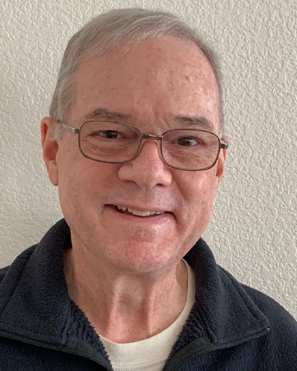 An older white man with white hair and metallic rimmed glasses in a white shirt and blue sweater smiles in front of a blank white wall
