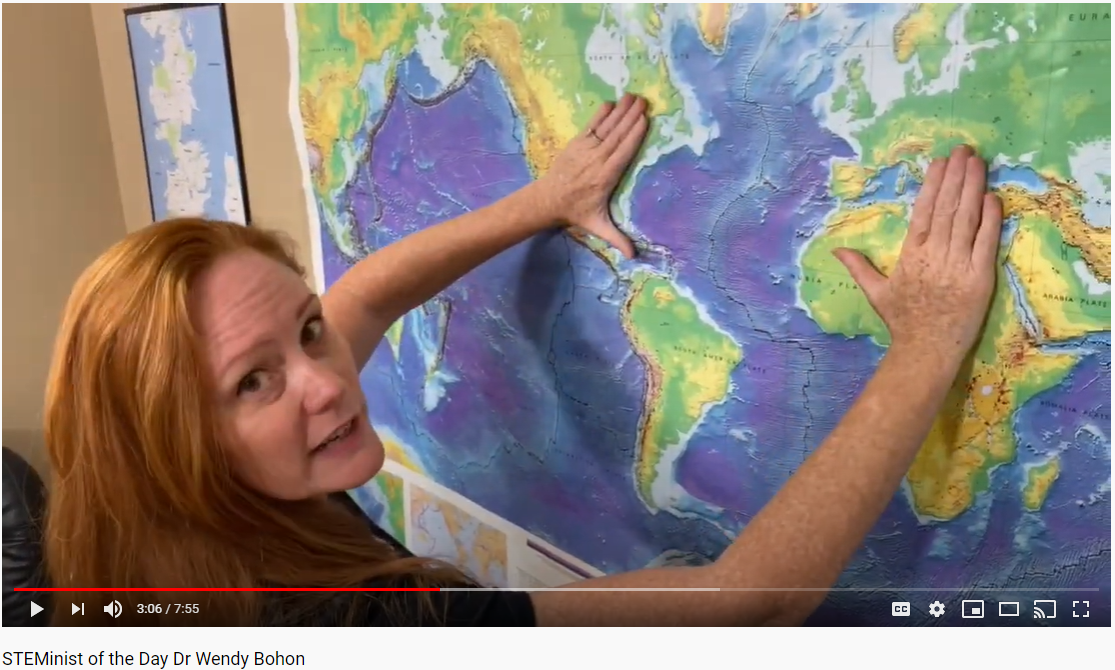 A screenshot of a smiling red-haired Caucasian woman in a black t-shirt who is gesturing to a world topographic map on the wall behind her. 