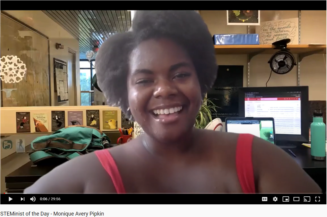 A screenshot of a smiling African American woman in a pink tanktop with her office in the background.