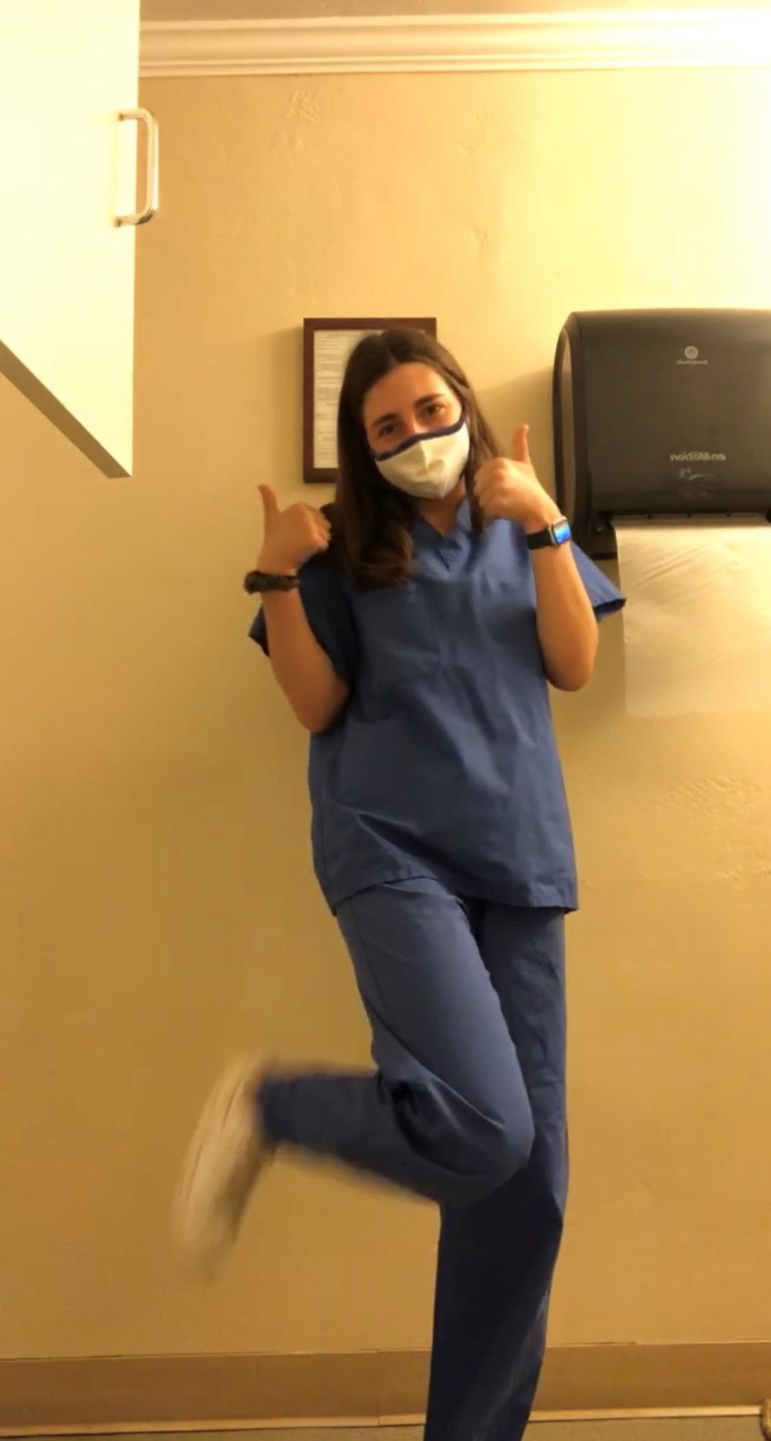 Image courtesy E. Lewis – Ellie in medical scrubs. A person standing in a medical office with long brown hair wears blue medical scrubs, a white facemask, and white sneakers. They give the camera two thumbs-up.