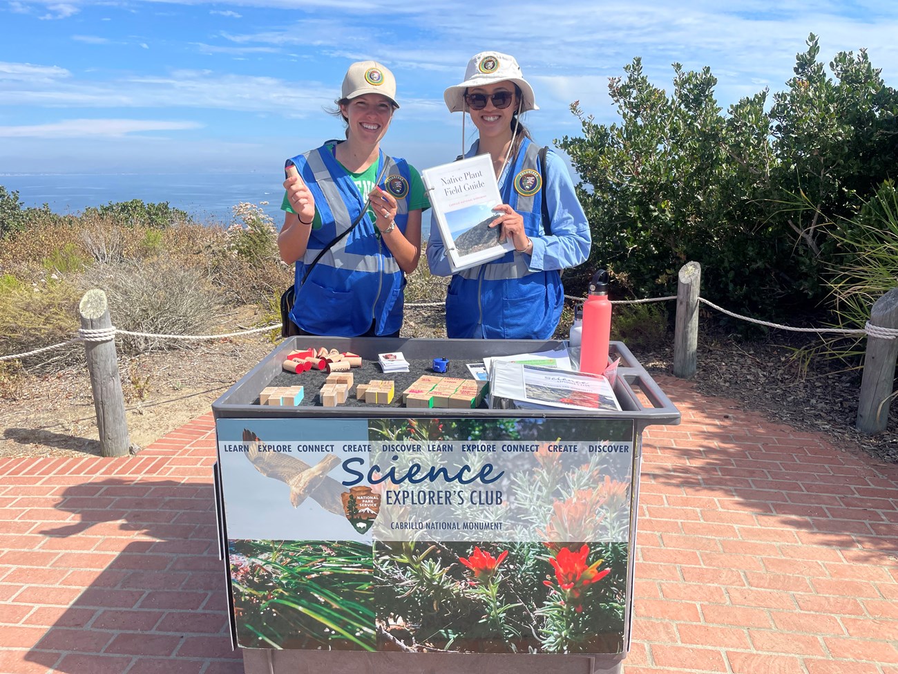 Two Science Explorer’s Club volunteers in bright blue vests stand behind the activity cart, which is covered in wooden blocks for the native plant activity. They are outdoors on a sunny day.