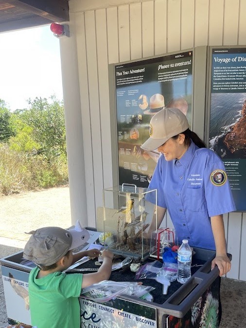 A Science Explorer’s Club volunteer in a blue shirt talks to a small child who is looking at the bird puzzle pieces for the bird physiology activity. They are in the Visitor Center walkway.