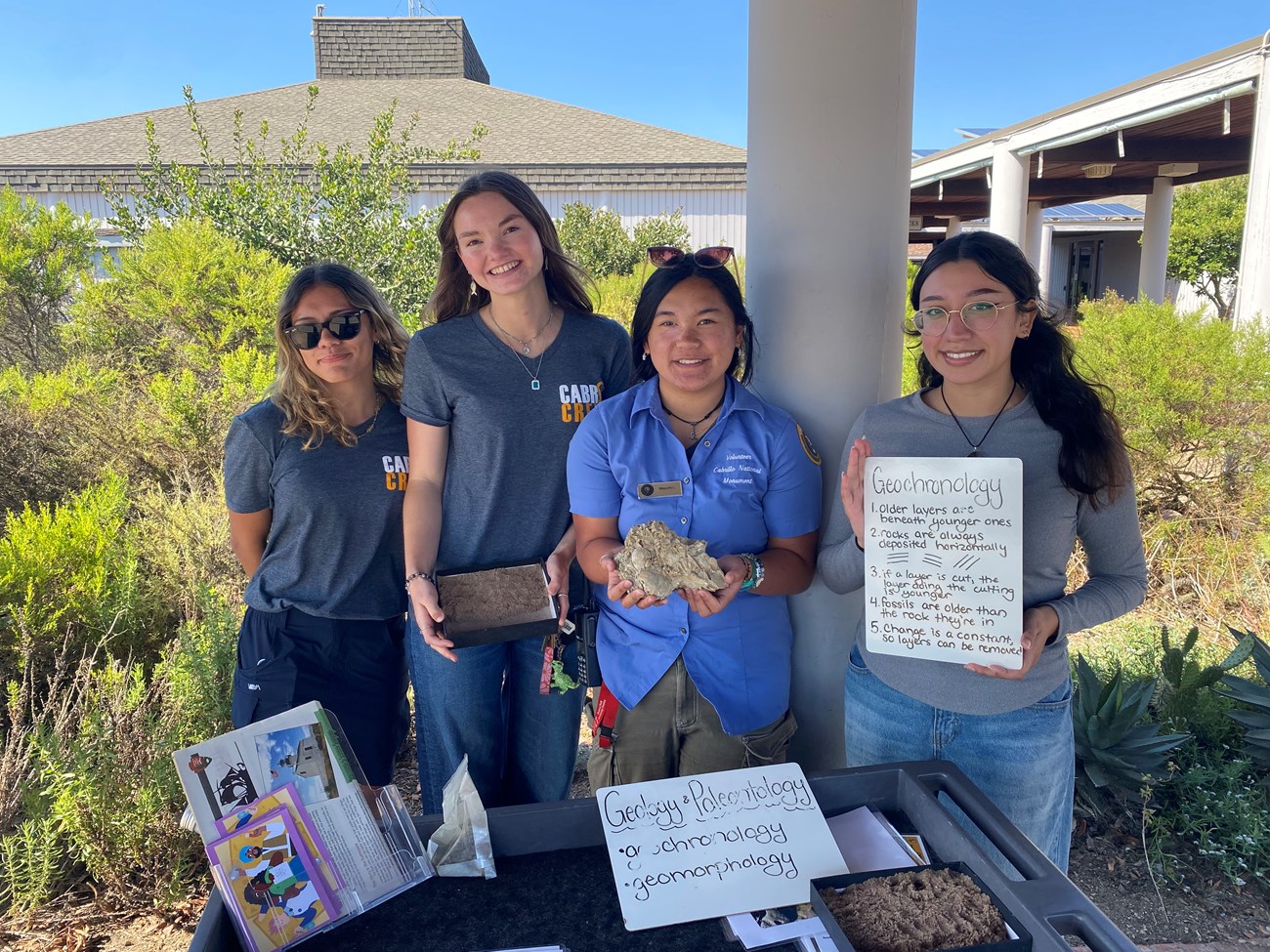 A group of four Science Explorer’s Club volunteers hold up rocks, fossils, and signs from the geology and paleontology activity. Behind them are the native plants around the Visitor Center.