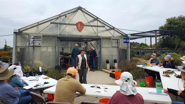 Volunteers sit behind white tables covered in native plant guides and garden tools. In front is a large glass building of the National Park Service Greenhouse.