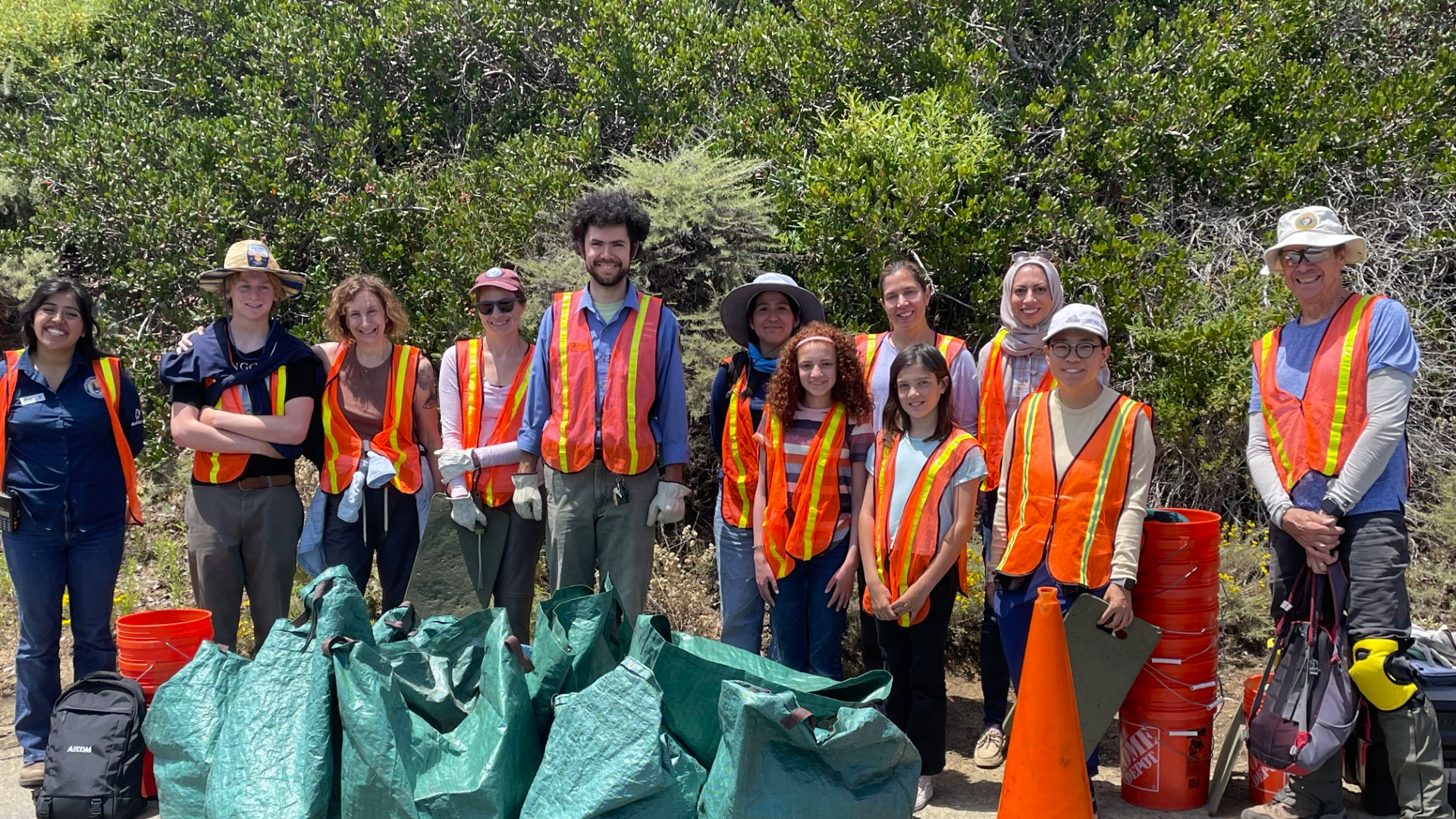 A group of volunteers from mixed age, gender, and backgrounds are smiling at the camera. They all wear orange safety vests. Behind them is the green native brush, and in front are the orange buckets, safety cones, and green bags full of plants.