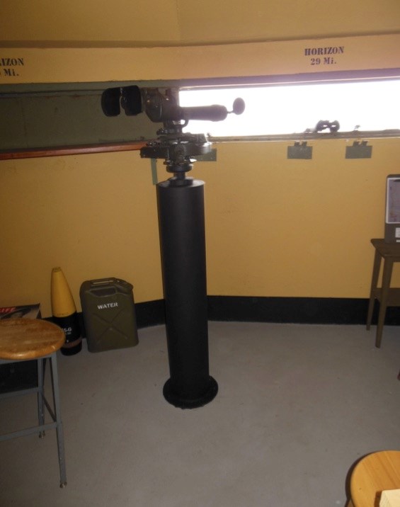 A WWII-era M1910A1 azimuth scope on a pedestal inside a bunker, positioned near a narrow observation window. The horizon is marked at 29 miles. Nearby are a water canister and a replica artillery shell.