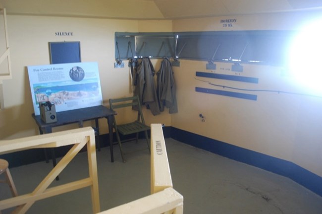 A partially restored WWII fire control room with a desk, informational signage, and coats hanging near observation slits. The wall shows distance markers to the horizon, and the space reflects the ongoing restoration of the lower bunker.