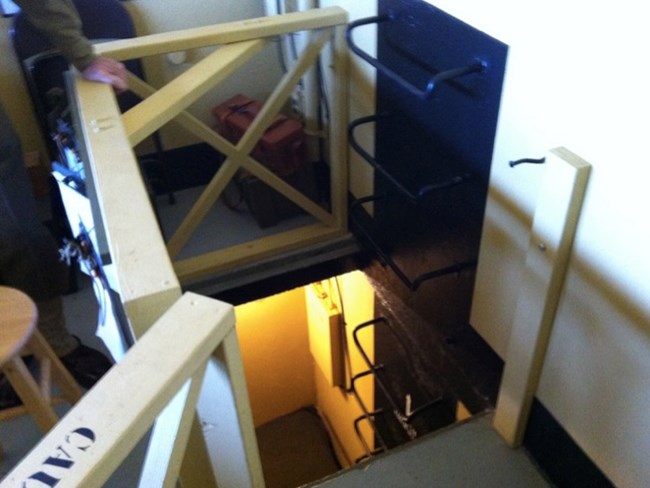 A view of the upper-level stairwell and ladder system leading to the lower bunker, with a restored wooden gate and black-painted ladder. A light illuminates the lower bunkroom, highlighting the compact design of the WWII-era bunker.