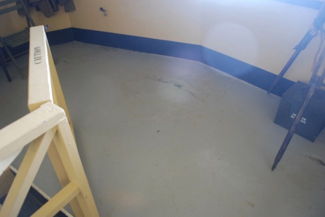 A bare section of the floor inside the bunker, showing worn marks where the M1910A1 azimuth scope was once mounted. The area is near a window, and a tripod is positioned nearby, giving context to the historical equipment once in place.