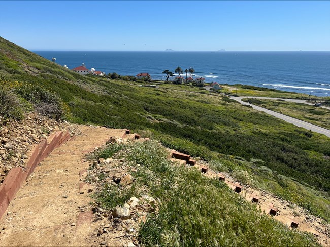 A right hand turn along a dirt trail leads to stairs.Low vegetation surrounds the trail. The blue ocean is seen in the distance.