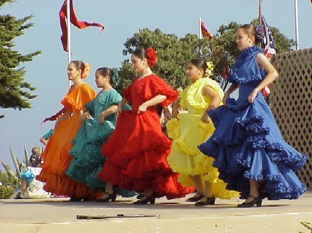 A group of six dancers, each dressed in brightly colored traditional Spanish dresses, perform on an outdoor stage. The dresses are in a variety of colors, including orange, green, red, yellow, and blue.