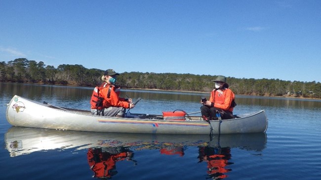 Two volunteers wearing reddish orange cold water protective gear are sitting in a canoe facing each other in the middle of a lake on a calm sunny day.