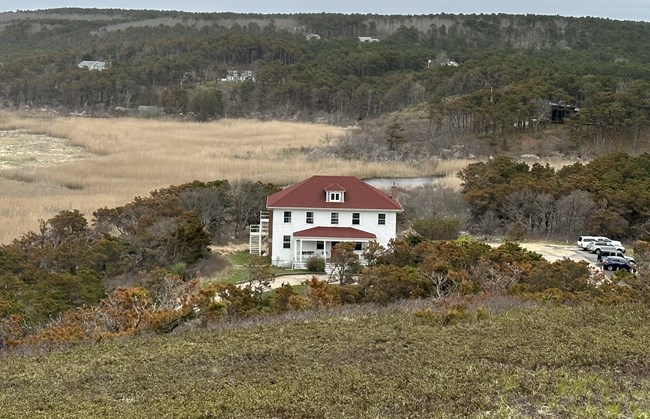 Two story white retangular building surrounded by mostly cedar trees adjacent to a salt marsh river valley