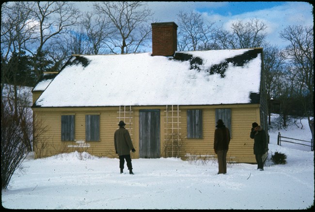 Three men stand around a small, one-story yellow house in a photo from the 1960's.