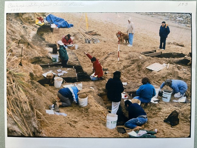Multiple individuals work on an archaeological excavation on a beach.