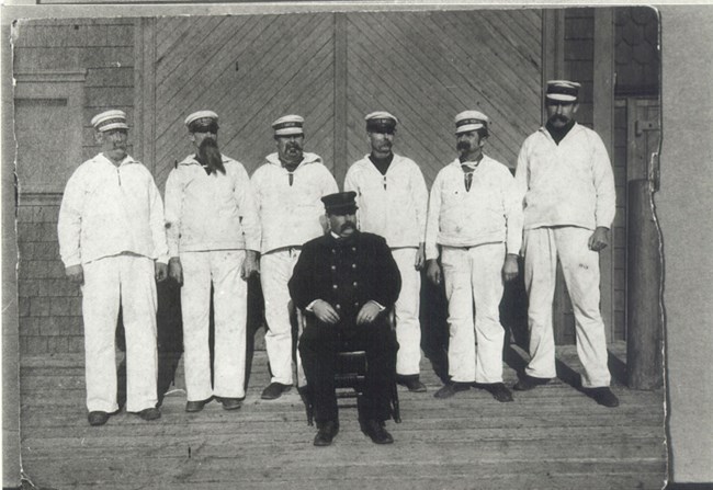 A group of six men stand in white with sailor's caps on and a man in black sits in front of them.