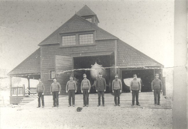 A group of men stand in front a building with an opening revealing a boat inside.
