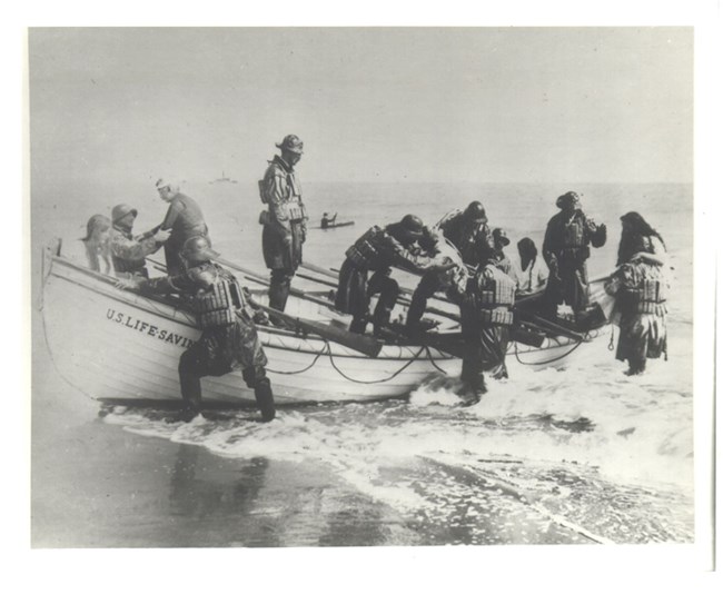 Photograph shows a boat beached on shore at the edge of the sea with people wearing cork lifevests surrounding the boat.