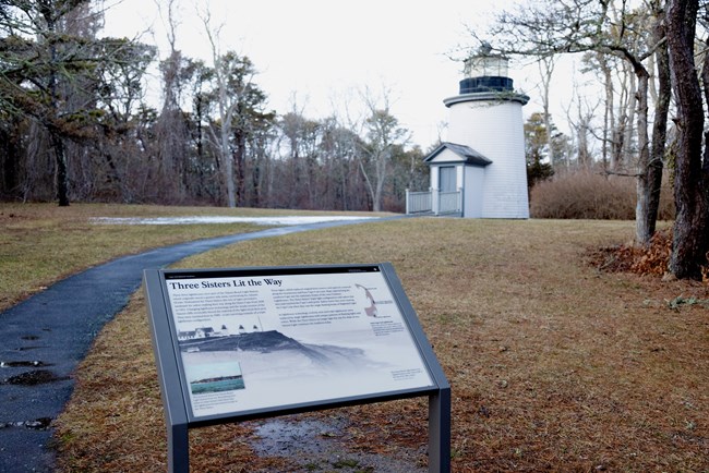 A sign reads "Three Sisters Lit the Way" and a short, white lighthouse can be seen to the right.