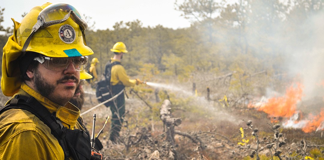 A man wearing yellow fire fighting equipment and safety glasses looks on as another person hoses down the fire with a water hose.