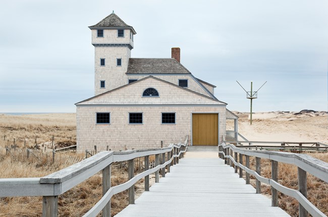 Boardwalk leads down to cedar-shingled building with a porch and tower.
