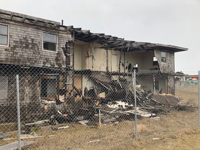 A dilapidated building sits behind a fence, with the walls removed and interior rooms exposed.