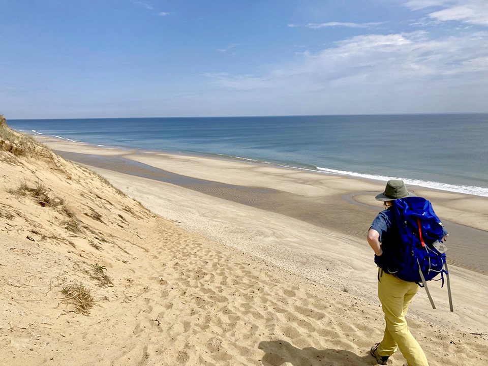A woman walks along a beach dune with the sea visible to the right.