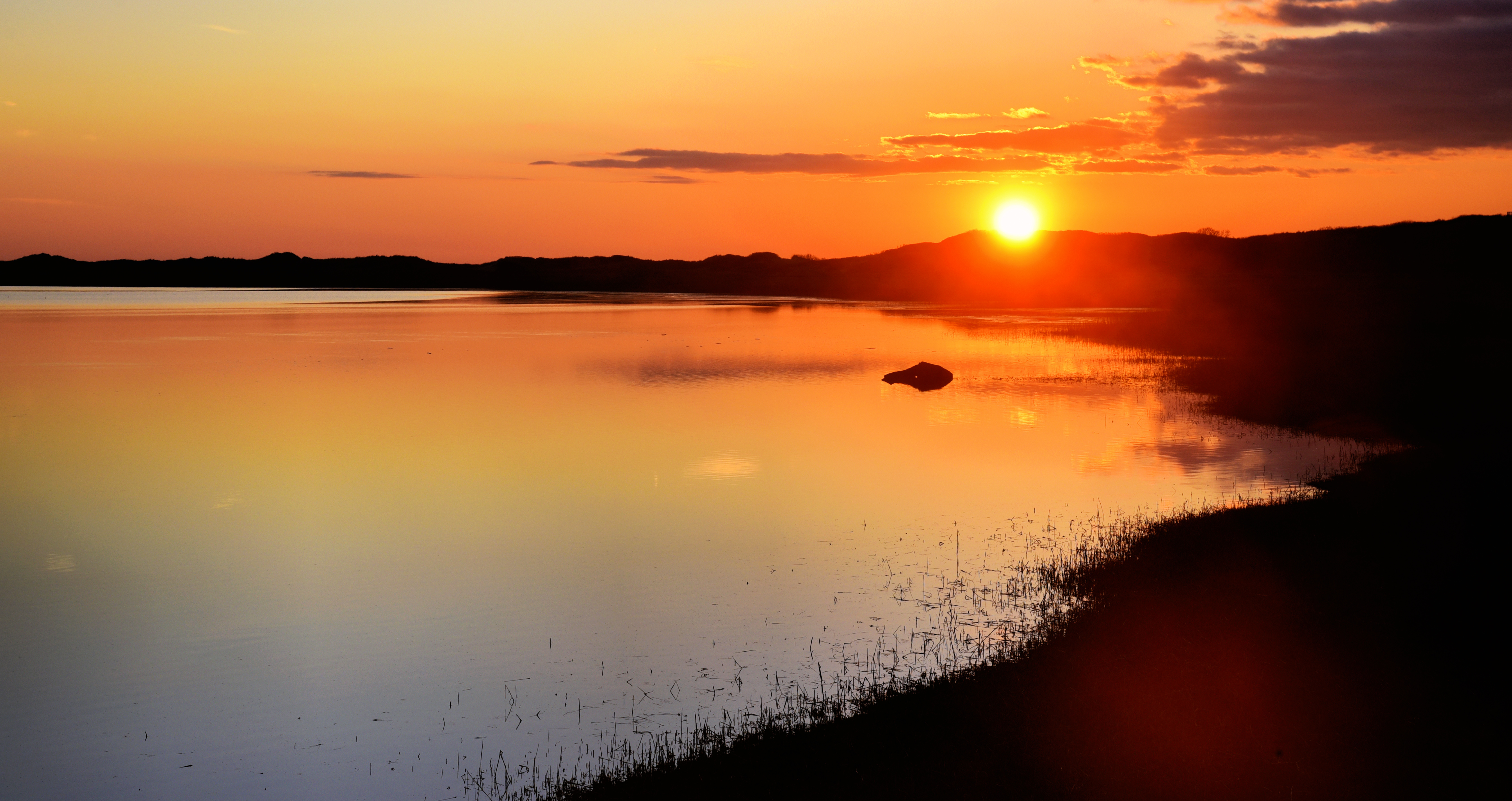 The sun sets over the Herring River and its banks with vibrant red, orange, pink, and blue colors in the sky reflecting in the water.