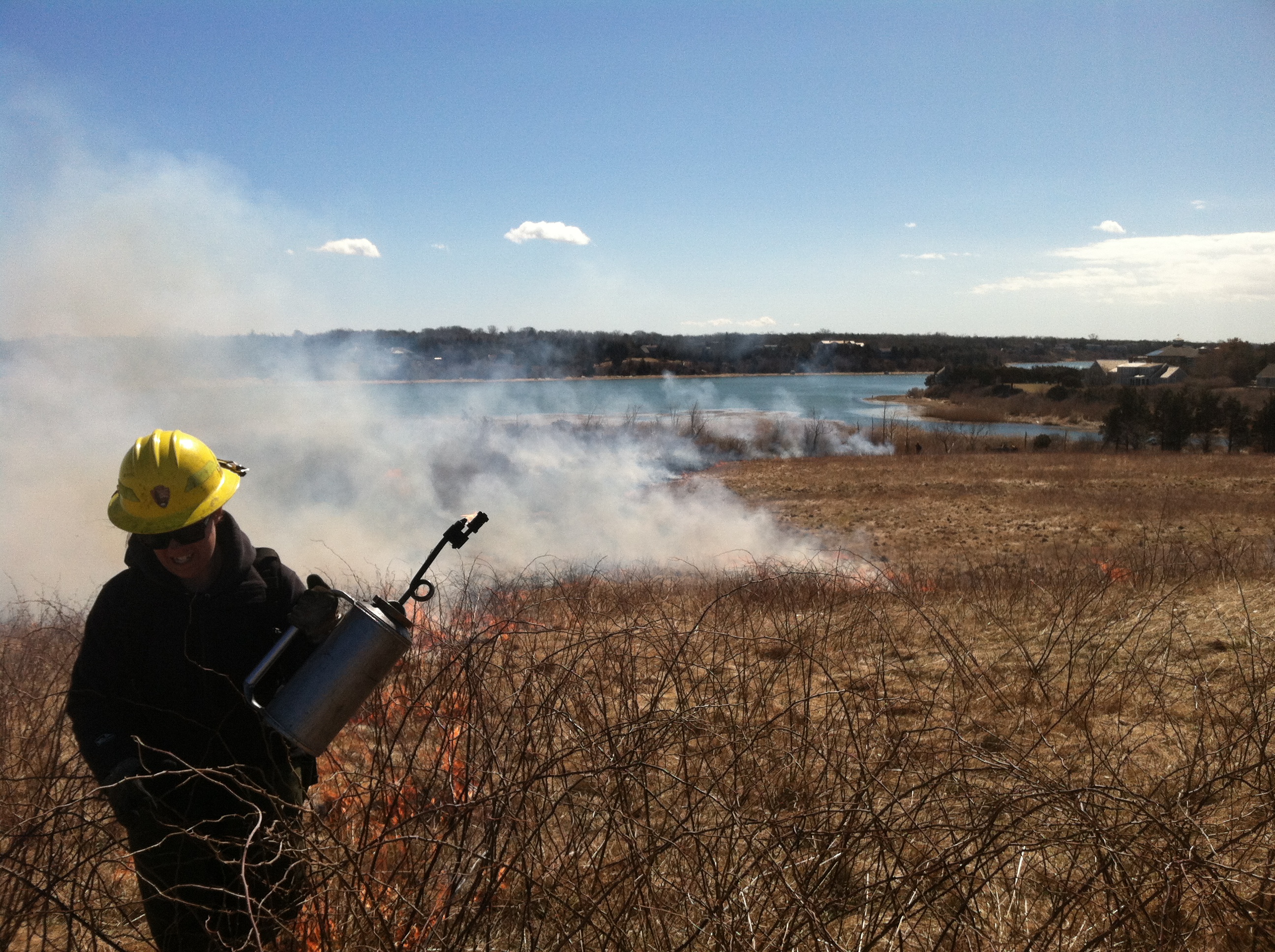 person in yellow hard hat holding a mental gas can walks along a meadow